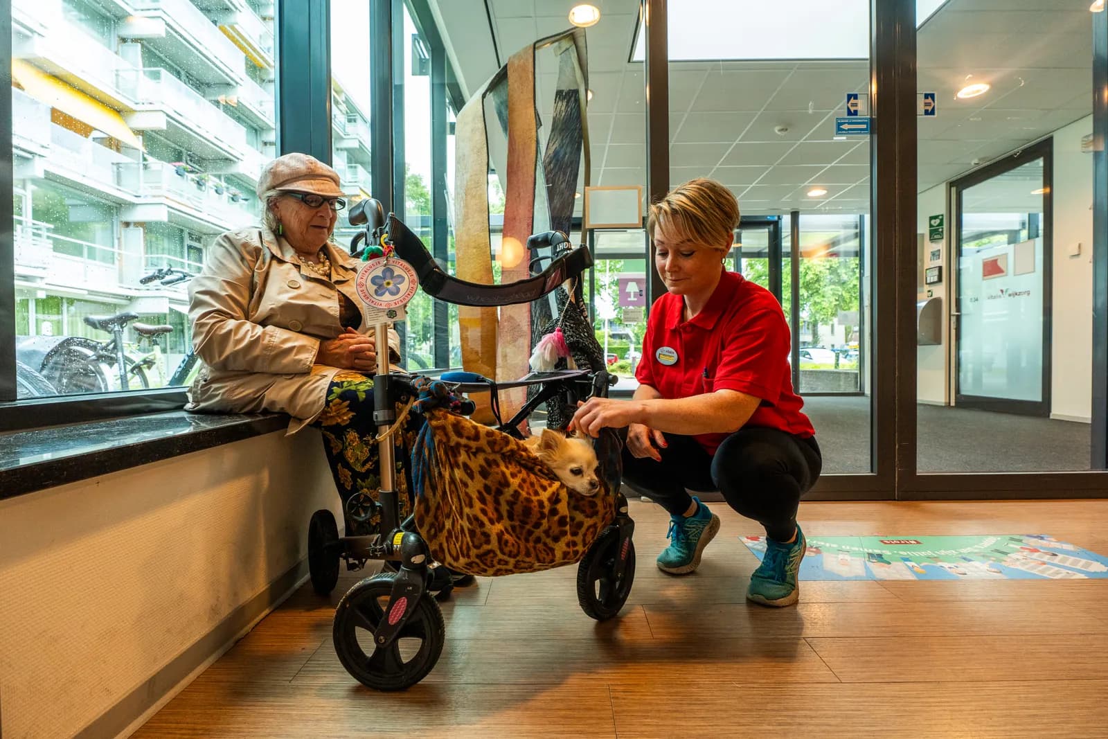 Een oudere met een rollator met daarin een hondje en een zorgmedewerker zijn aan het kletsen